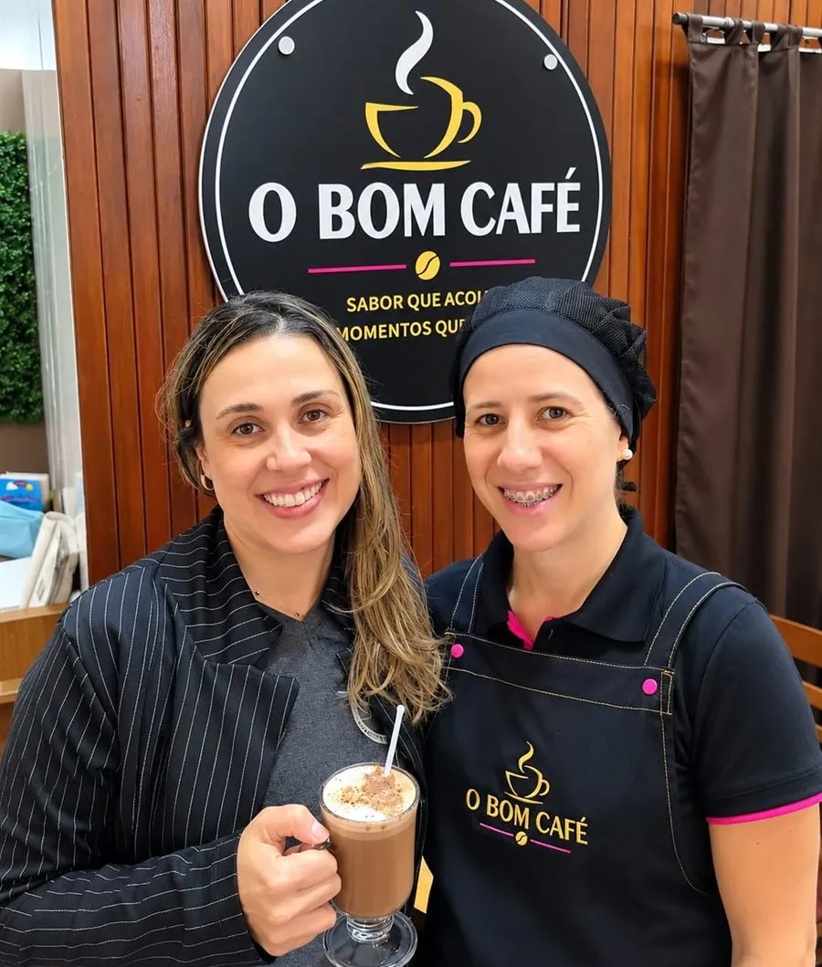 Duas integrantes da equipe do O Bom Café sorrindo em frente à logo, uma delas de avental