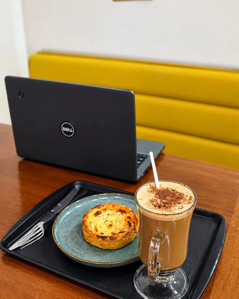 Notebook, café com chantili e uma quiche sobre a mesa, com o sofá amarelo ao fundo