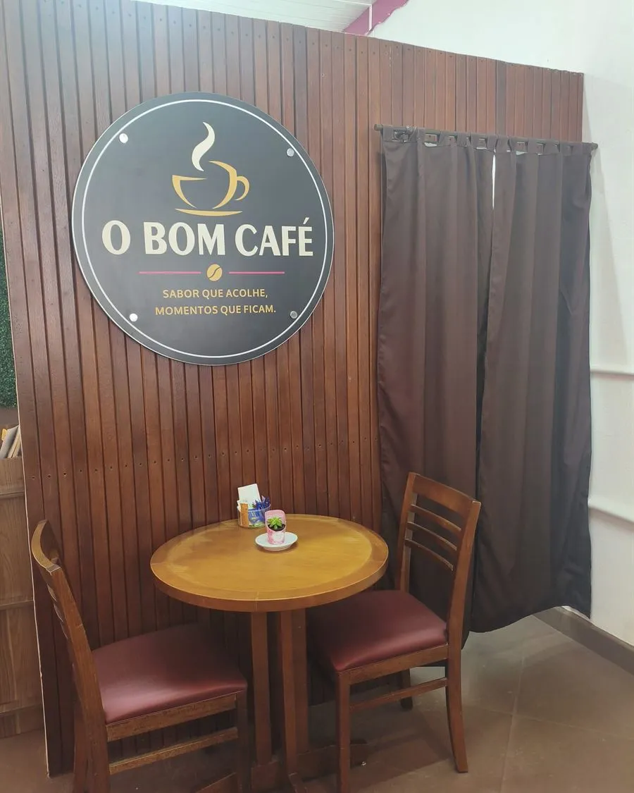 Cantinho do O Bom Café: mesa redonda e cadeiras de madeira em frente à parede com a logo